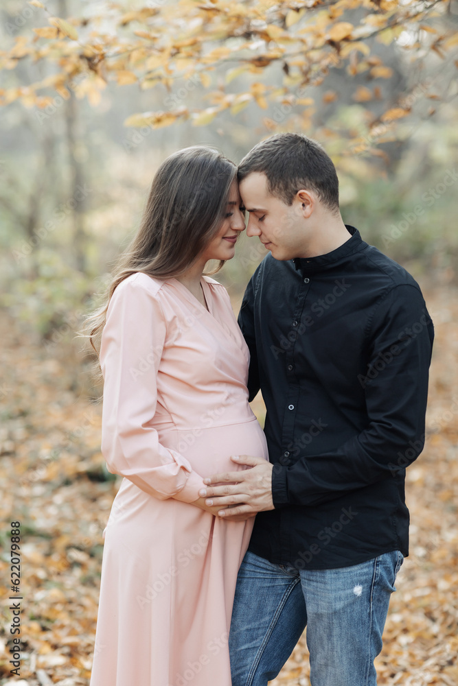 portrait of a happy young family, husband and pregnant wife, standing against the background of beautiful light in the autumn forest, looking at each other portrait of a happy young family, husband and pregnant wife, standing against the background of beautiful light in the autumn forest, looking at each other
