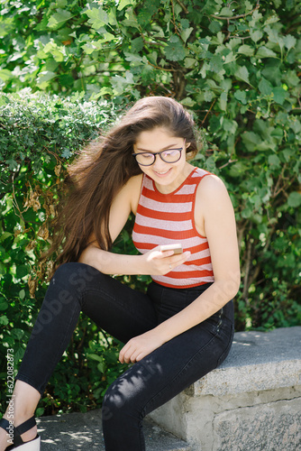 Smiling Young Asian woman with glasses using smartphone and sending sms. Young woman on city background with green leaves.