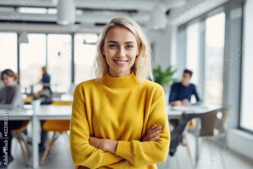 Generative AI image of portrait of smiling young female employee in sweater looking at camera while standing with hands crossed in blurred and illuminated office with colleagues