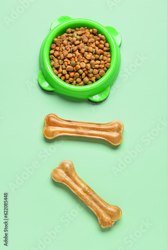 Bowl with dry dog food and chew bones on color background