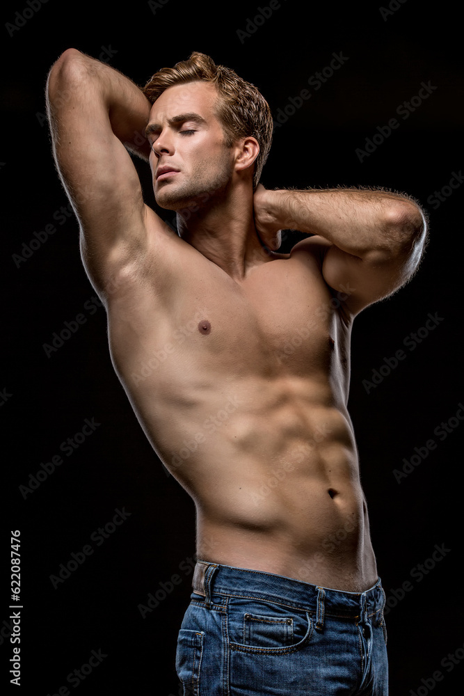 Strong man in blue jeans with naked torso stands with closed eyes on the black background in the studio. He holds his hands on the neck behind the head. Vertical low-key photo.