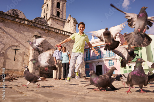 Happy tourist on holidays during vacation trip. Hispanic people traveling in Havana, Cuba. Grandpa and grandson feeding birds, with child running chasing pigeons © Designpics