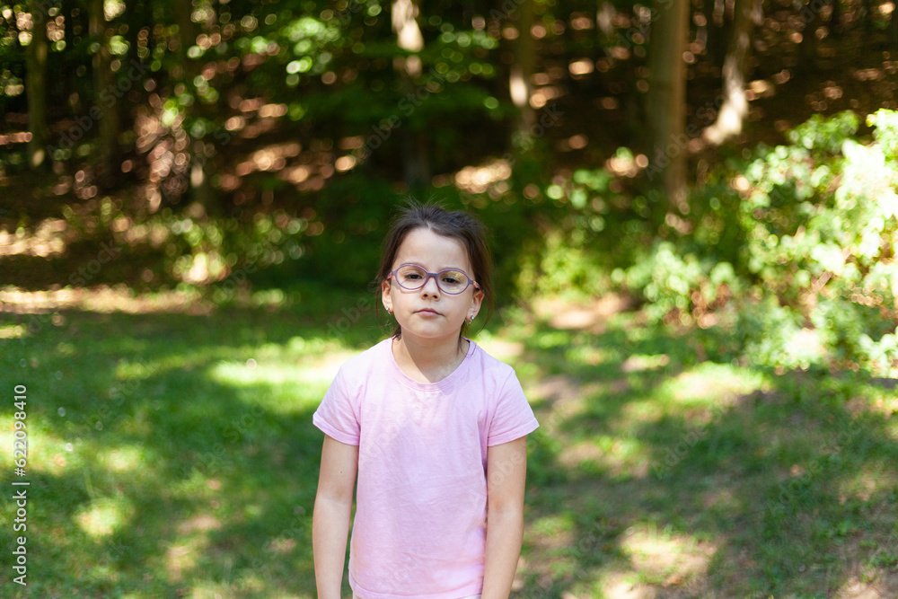 Little girl in glasses and pink t-shirt standing in the park Little girl in glasses and pink t-shirt standing in the park