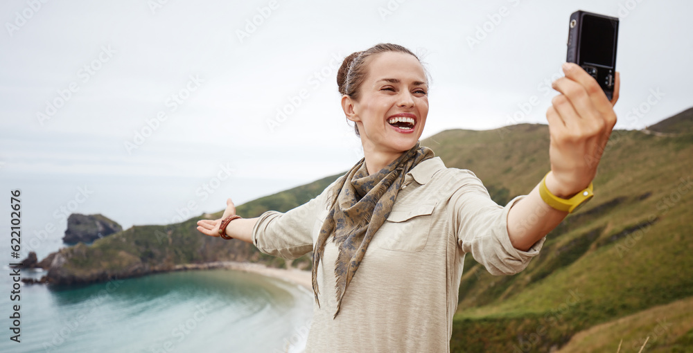 Into the wild in Spain. smiling active woman hiker taking selfie with digital camera in front of ocean view landscape