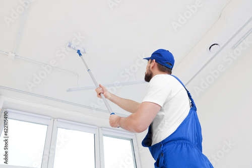 Worker in uniform painting ceiling with roller on stepladder indoors, low angle view