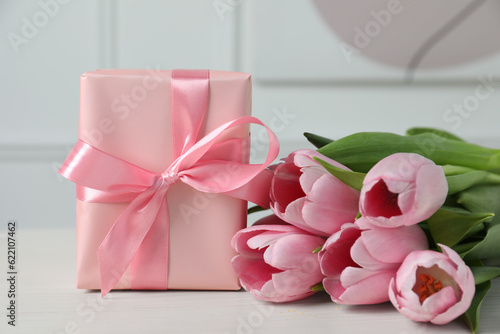 Beautiful gift box with bow and pink tulips on white table, closeup