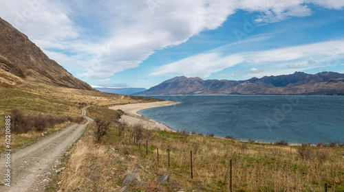 Narrow gravel road along a lake shore with mountains on background. Lake Hawea, New Zealand