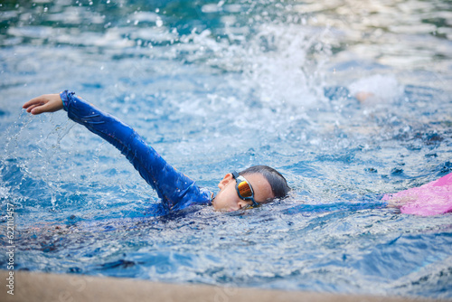 Cute little Asian 7 years old school boy child in swimming goggles and swimming suit learning to swim at outdoor pool in summer day, Little kid uses a foam pad to practice swimming in a swimming pool