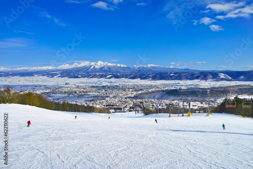 快晴の富良野スキー場から見る十勝岳連峰、北の峰ゾーンの広大な初心者コースを滑るスキーヤー達 