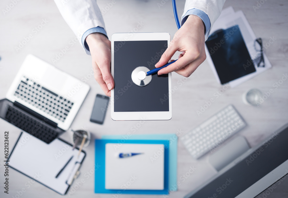 Doctor checking heartbeat on a digital tablet using a stethoscope, desktop with medical equipment on background, top view Doctor checking heartbeat on a digital tablet using a stethoscope, desktop with medical equipment on background, top view
