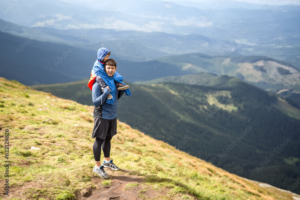 Father in sportswear stands on the mountain and holds his son on the shoulders on the background of the mountains and the cloudy sky. Boy wears multi-colored big size windcheater. Horizontal.