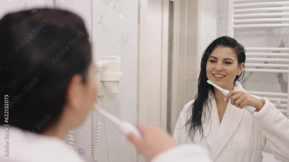 Brushing teeth, Mirror Reflection, Bathroom Activity. Appealing adult woman brushing teeth post-shower, gazing at mirror in bathroom, donned in white silky bathrobe Brushing teeth, Mirror Reflection, Bathroom Activity. Appealing adult woman brushing teeth post-shower, gazing at mirror in bathroom, donned in white silky bathrobe