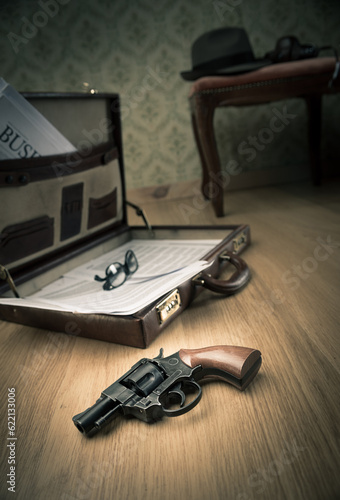 Detective open briefcase with vintage gun on the floor and borsalino hat on background.