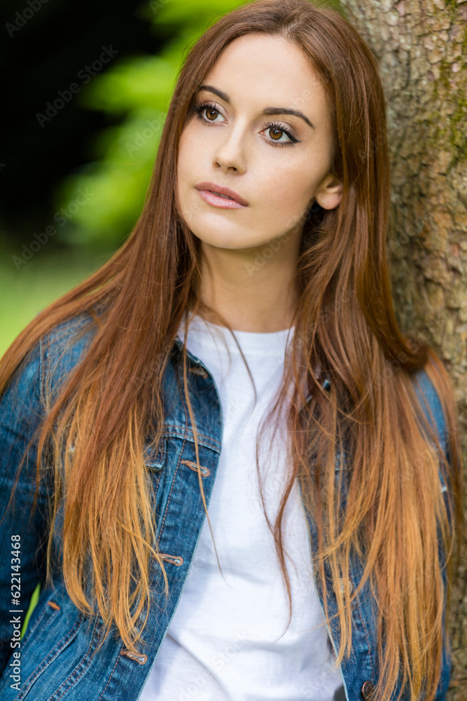Outdoor portrait of beautiful thoughtful sad girl or young woman with red hair wearing a white t-shirt and denim jeans jacket leaning against a tree in the countryside Outdoor portrait of beautiful thoughtful sad girl or young woman with red hair wearing a white t-shirt and denim jeans jacket leaning against a tree in the countryside