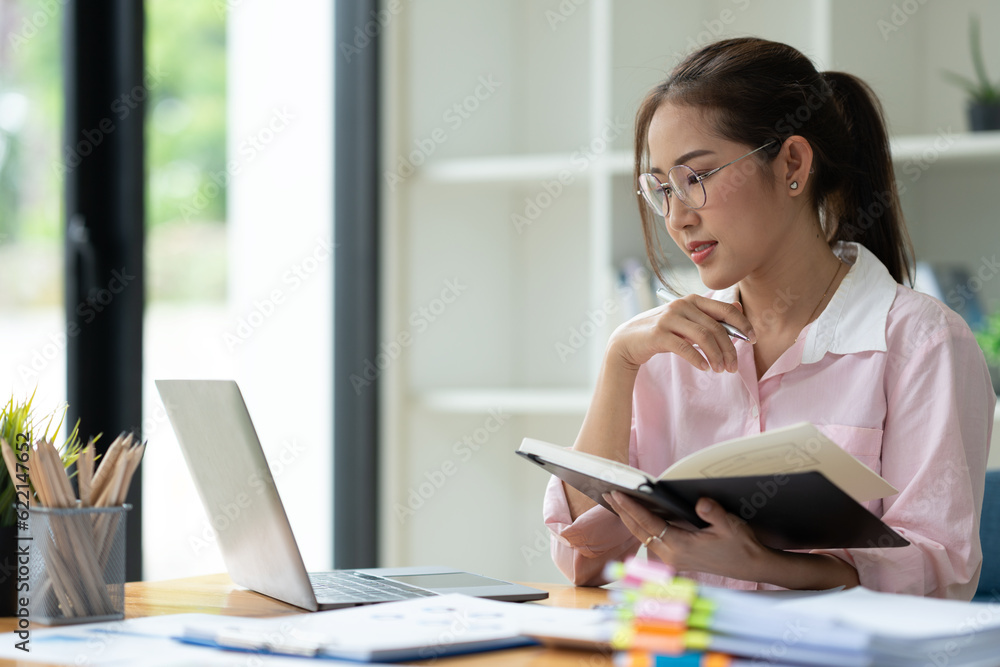 Office worker or businesswoman, attractive Asian woman in eyeglasses researching Analyze information about the business plan. take notes in a book Notebook while using a laptop computer at her desk. Office worker or businesswoman, attractive Asian woman in eyeglasses researching Analyze information about the business plan. take notes in a book Notebook while using a laptop computer at her desk.