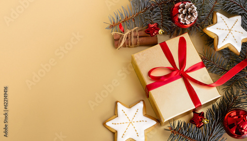 Christmas decoration composition on light gold background with beautiful Golden gift box with red ribbon, fir branches, cones, stars, Christmas cookies, cinnamon, top view, copy space, banner format. © Uuganbayar