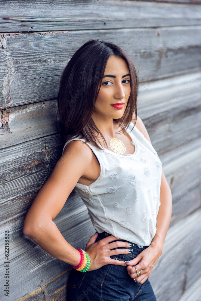 Fototapeta premium Girl brunette in a park in the summer resting white blouse on the background of a wooden fence, fashion lifestyle, gentle look. Sensual make-up, tanned skin and bright red lipstick. Portrait of a woma