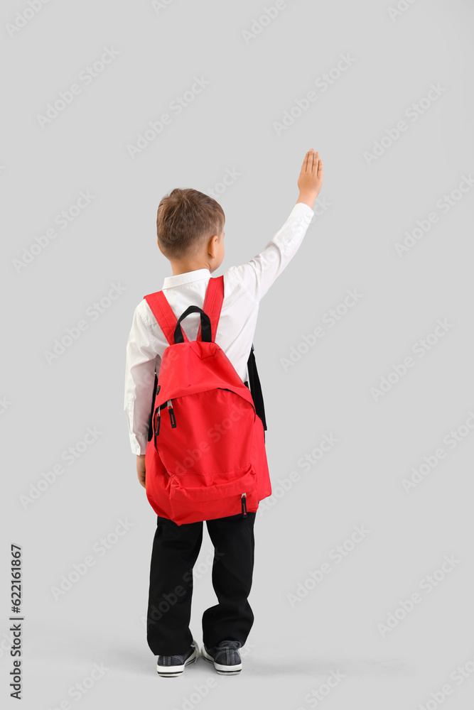 Little schoolboy with red backpack waving hand on grey background, back view Little schoolboy with red backpack waving hand on grey background, back view