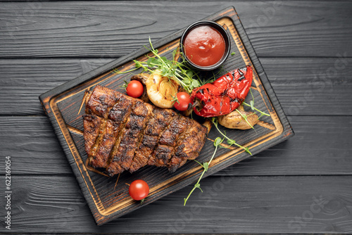 Grilled pork bbq ribs served with cherry tomatoes, pepper and barbeque sauce on wooden cutting board over dark background. Top view