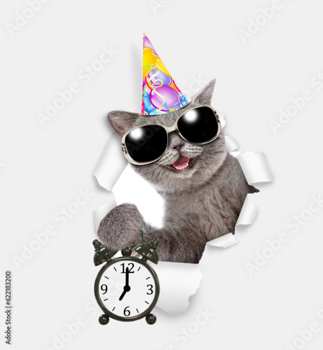 Happy cat wearing sunglasses and party cap looks through a hole in white paper and shows alarm clock © Ermolaev Alexandr