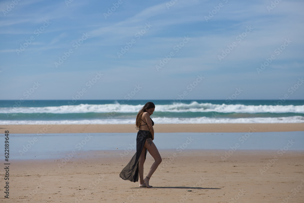 Beautiful young woman with long brown hair in swimming costume and black silk sarong strolls along the shore of the beach enjoying the peace and quiet of the sunny day. In the background the blue sea. Beautiful young woman with long brown hair in swimming costume and black silk sarong strolls along the shore of the beach enjoying the peace and quiet of the sunny day. In the background the blue sea.