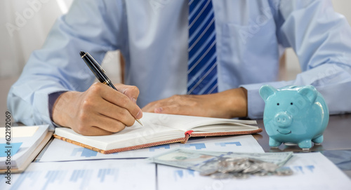 Businessman hand writing income and expenses to account with piggy bank and dollar notes, step up start-up businesses to success, Saving money for future plan and retirement fund concept