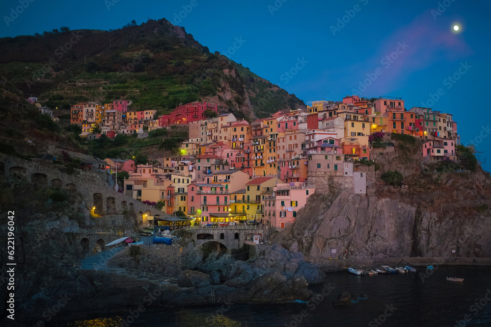 Cinque Terre, Italy - night view of the colorful pastel houses in Manarola, a seaside village on the rugged Italian Riviera coast. Moon in sky, boats in water. Summer travel vacation background.
