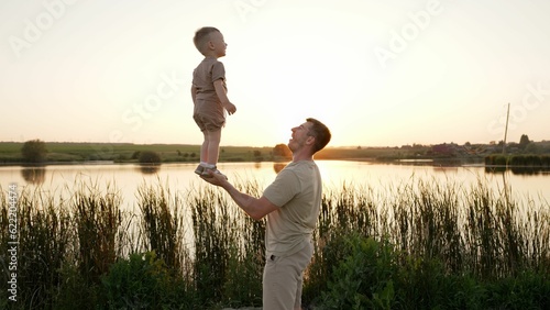 The trust of the son to the father. The father holds his son in full growth in the palm of his hand. Dad and son doing gymnastic exercises in nature. The father holds his son in one hand.