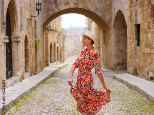 summer trip to Rhodes island, Greece. Young Asian woman in ethnic red dress walks Street of Knights of Fortifications castle. female traveler visiting southern Europe. Unesco world heritage site. © YURII Seleznov