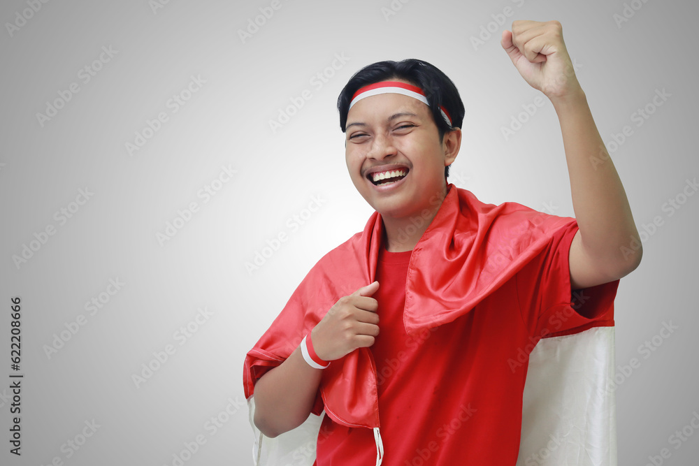 Portrait of attractive Asian man in t-shirt with red white ribbon on head with flag on his shoulder as a cloak, raising his fist, celebrating Indonesia's independence day. Isolated image on gray Portrait of attractive Asian man in t-shirt with red white ribbon on head with flag on his shoulder as a cloak, raising his fist, celebrating Indonesia's independence day. Isolated image on gray