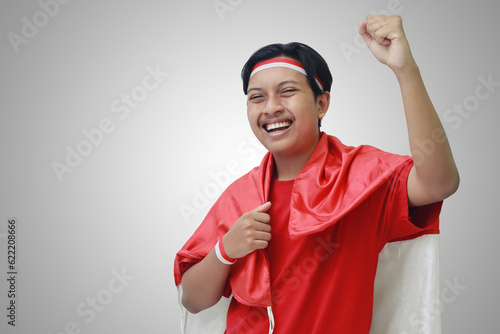 Portrait of attractive Asian man in t-shirt with red white ribbon on head with flag on his shoulder as a cloak, raising his fist, celebrating Indonesia's independence day. Isolated image on gray