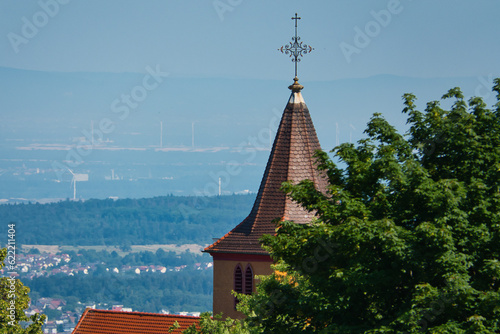 Eine Kirchturmspitze mit einem Kreuz aus Metall auf dem Dach, halb verdeckt von einem Baum, mit Blick über weites Land im Hintergrund