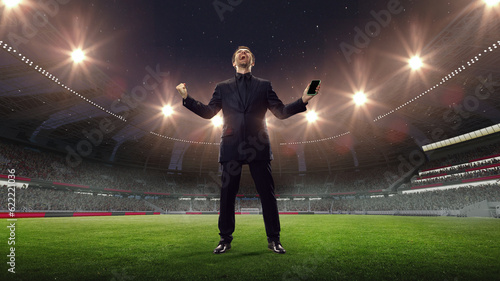 Happy, lucky, excited businessman standing with mobile phone, feeling success and win of money on sports betting. 3D arena background. Concept of sport, fan, betting, finances, gambling, bookmaker