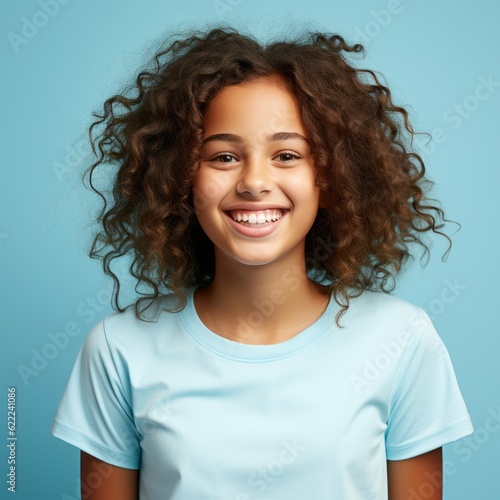 Portrait of a smiling African little girl with brown hair on a blue background. Happy small African kid with a smile and curly hair. Cheerful dark-skinned child in a blue shirt with shiny white teeth © Valua Vitaly