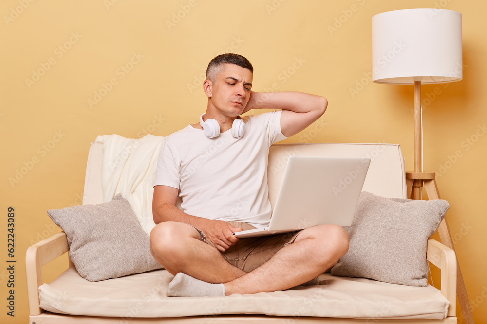 Internet connection. Notebook work. Laptop technology. Tired young man working on laptop computer feeling pain massaging painful neck sitting on comfortable sofa at home Internet connection. Notebook work. Laptop technology. Tired young man working on laptop computer feeling pain massaging painful neck sitting on comfortable sofa at home