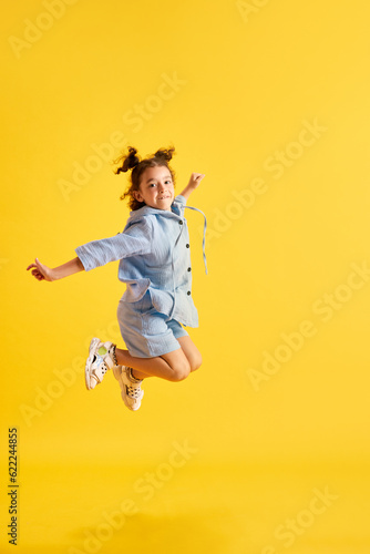 Full-length portrait of active little kid, girl, child in casual clothes cheerfully jumping against yellow studio background. Concept of emotions, childhood, education, fashion, lifestyle, ad