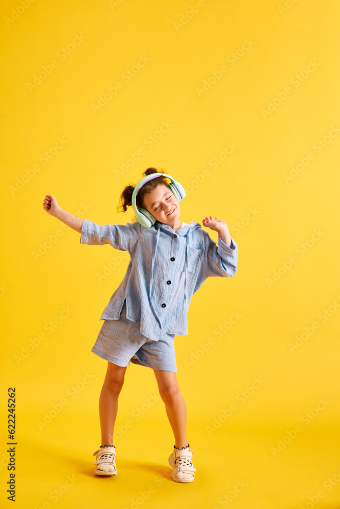 Full-length portrait of beautiful little child, girl listening to music in headphones and dancing against yellow studio background. Concept of emotions, childhood, education, fashion, lifestyle, ad Full-length portrait of beautiful little child, girl listening to music in headphones and dancing against yellow studio background. Concept of emotions, childhood, education, fashion, lifestyle, ad