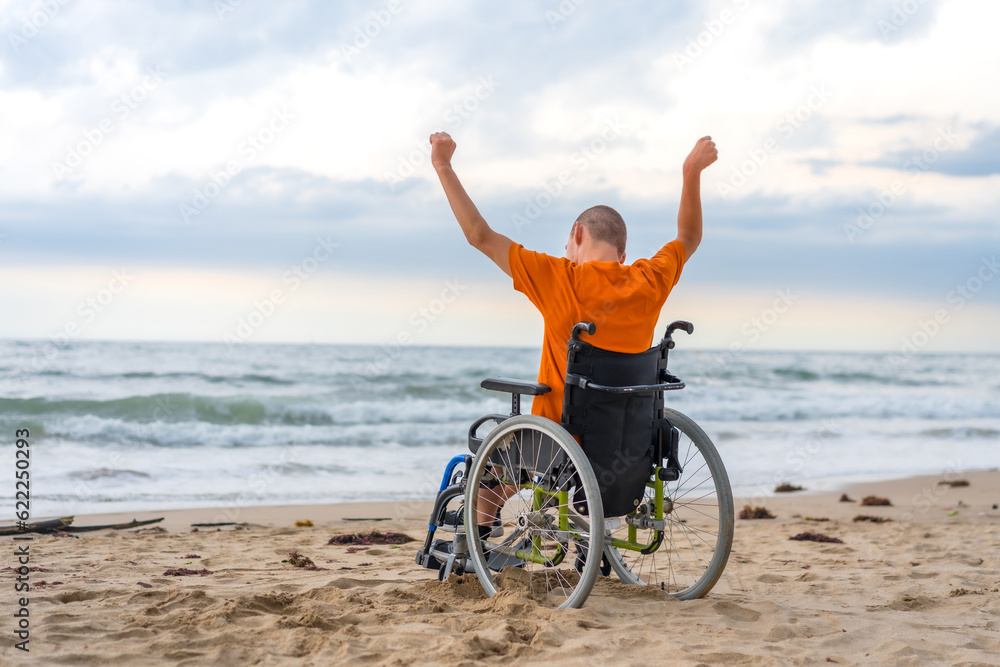 Fototapeta premium Disabled person on his back in a wheelchair on the beach with open arms enjoying the freedom