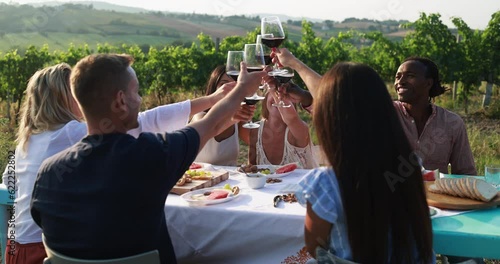 Happy adult friends having fun cheering with red wine and eating together outdoor - Vineyard in background