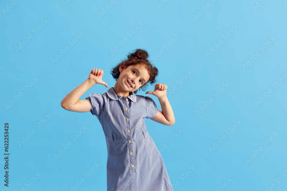 Portrait of lovely little girl, child with curly hair and cute hairdo posing against blue studio background. Concept of emotions, childhood, education, fashion, lifestyle, ad Portrait of lovely little girl, child with curly hair and cute hairdo posing against blue studio background. Concept of emotions, childhood, education, fashion, lifestyle, ad