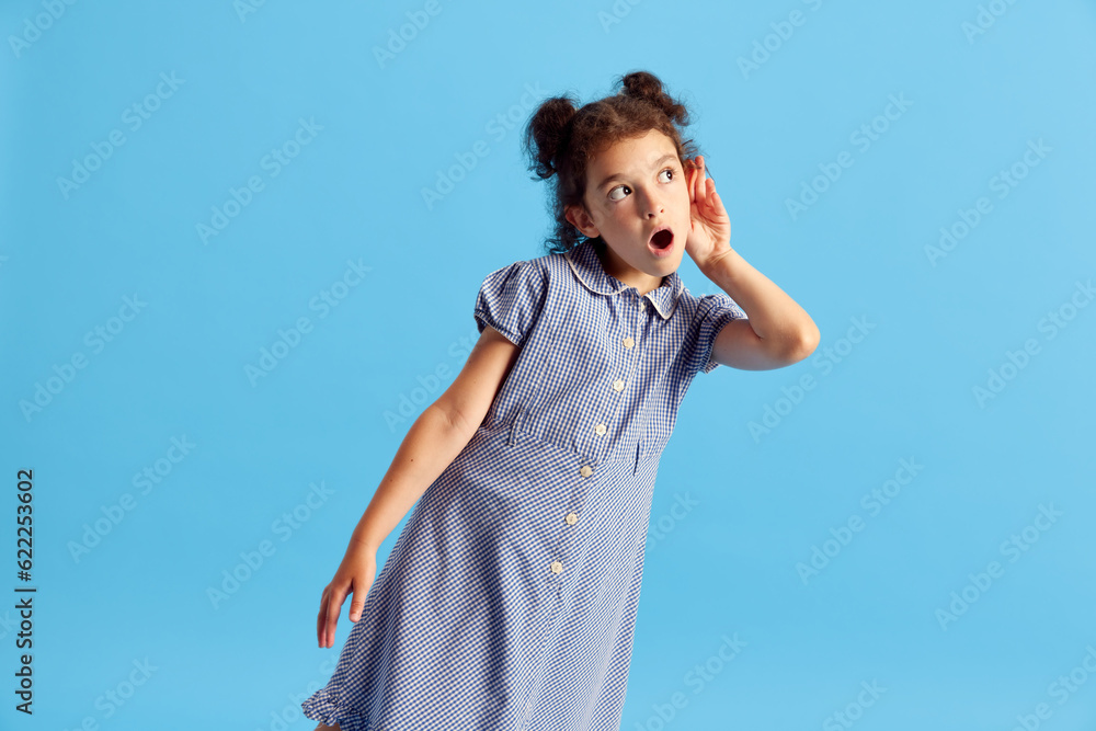 Portrait of lovely little girl, child with curly hair and cute hairdo posing against blue studio background. Overhearing. Concept of emotions, childhood, education, fashion, lifestyle, ad Portrait of lovely little girl, child with curly hair and cute hairdo posing against blue studio background. Overhearing. Concept of emotions, childhood, education, fashion, lifestyle, ad