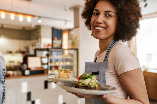 Cheerful waitress carrying plates with meal while working in cafe