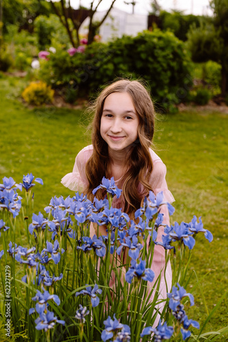 A little girl with long wavy brown hair stands and enjoys the smell of beautiful blue shrub flowers in the backyard of a house in the village on a flower bed