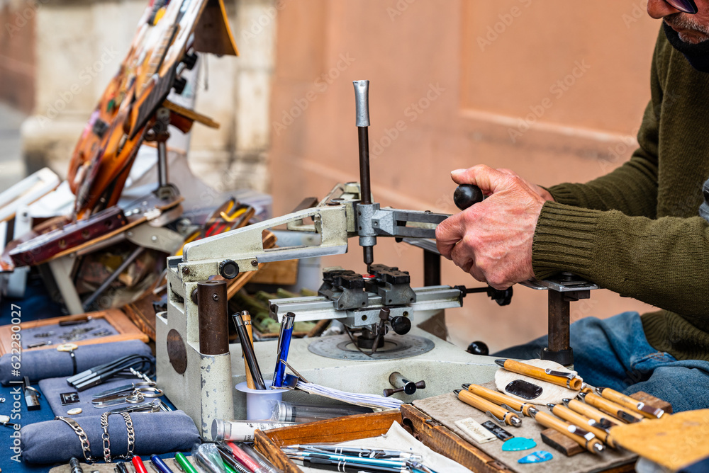 Elderly engraver at work on a street bench. Detail of the hands engrave plaques, pens and other objects. Old jobs. Elderly engraver at work on a street bench. Detail of the hands engrave plaques, pens and other objects. Old jobs.