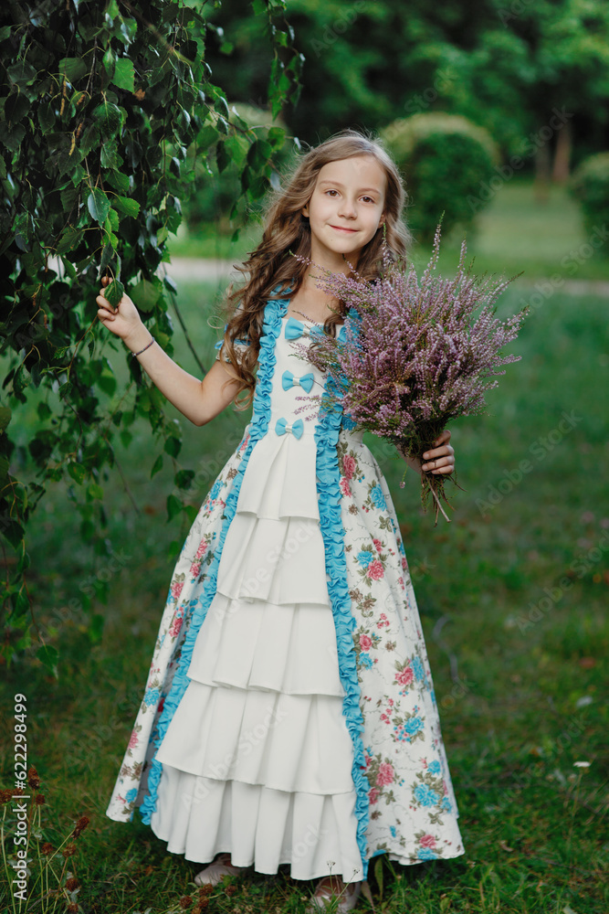 Pretty little smiling girl with bouquet of heather flowers in long vintage dress standing in summer park with green background, vertical lifestyle outdoor kid's portrait in provence country style