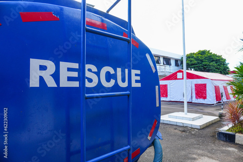 Selective focus on the 'Rescue' writing on the back of a blue water tank vehicle with emergency disaster tent in the background