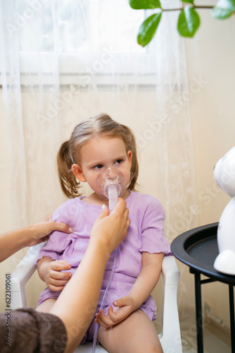 Cute little girl uses inhaler at home. Holding mask over her nose and mouth with help of her mother 