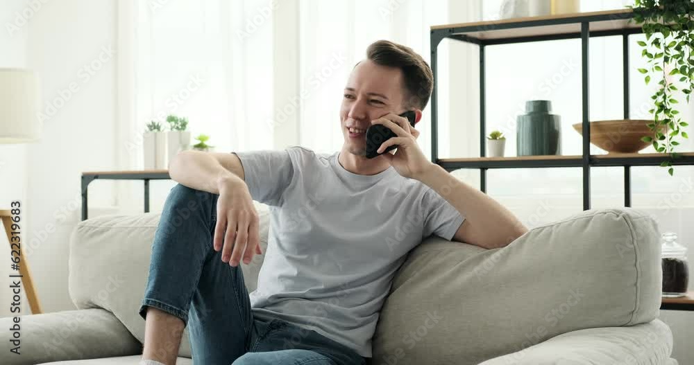 A joyful Caucasian man is sitting on a couch and engaging in a phone conversation. With a bright smile on his face, he expresses happiness and enthusiasm as he communicates. A joyful Caucasian man is sitting on a couch and engaging in a phone conversation. With a bright smile on his face, he expresses happiness and enthusiasm as he communicates.