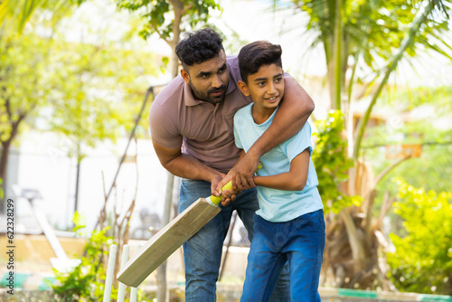 indian father teaching his son to playing cricket game at park - concept of Shared passion, family bonding and fatherhood