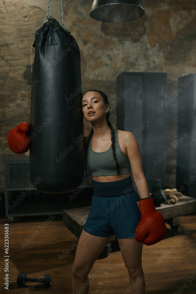 Young sportive woman boxer standing nearby punching bag at retro gym